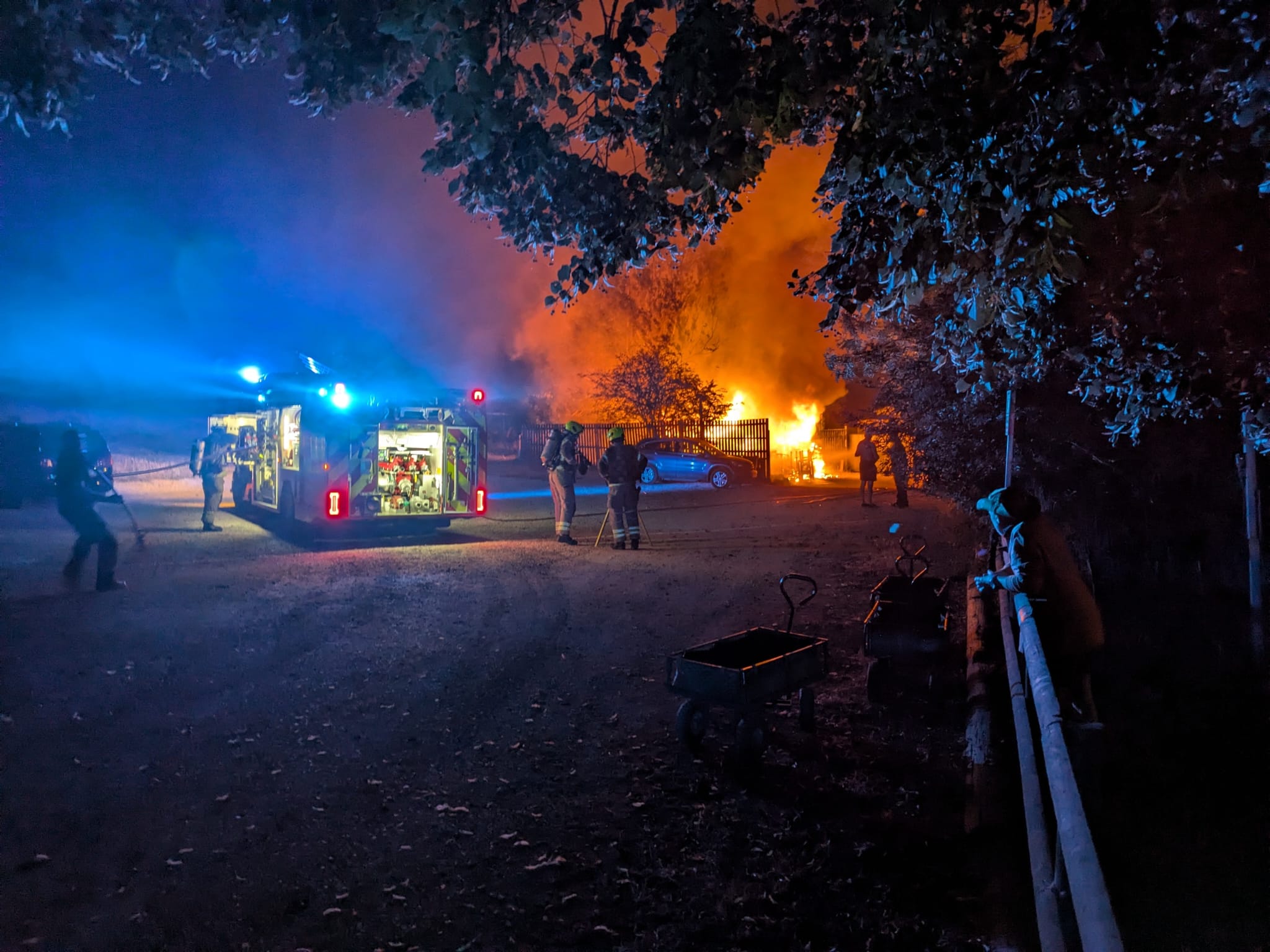 Full image of Firefighters pull hoses from a fire engine towards the inferno of a flaming cabin.