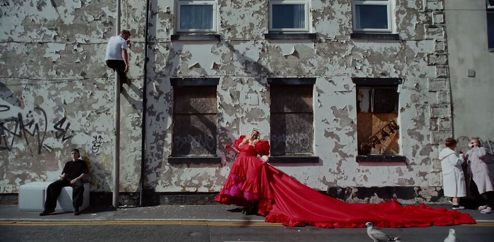 A partially-boarded up and heavily-graffitied residential street with unmaintained peeling paint, in front of which a woman in an enormous red dress walks.