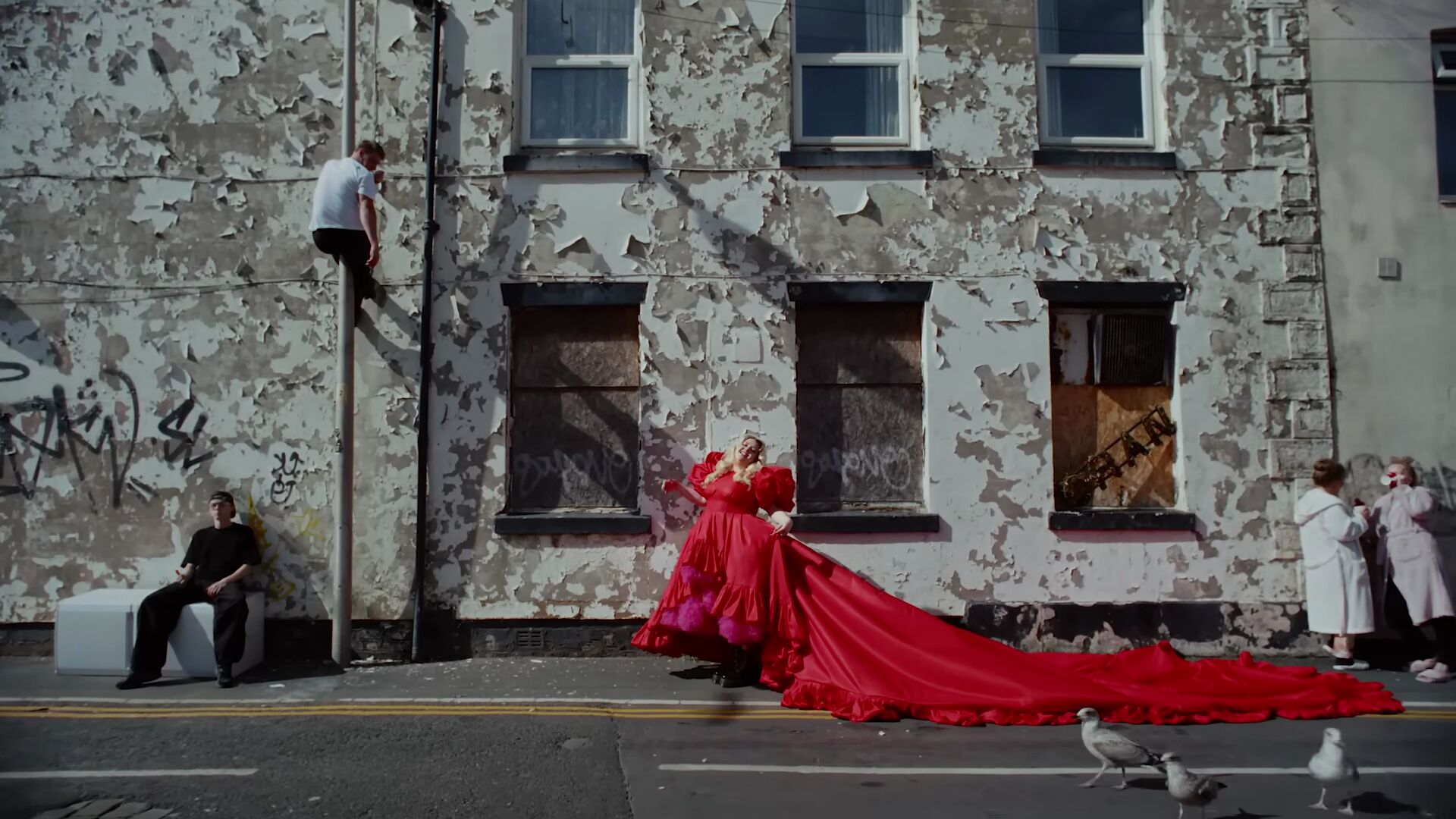 Full image of A partially-boarded up and heavily-graffitied residential street with unmaintained peeling paint, in front of which a woman in an enormous red dress walks.