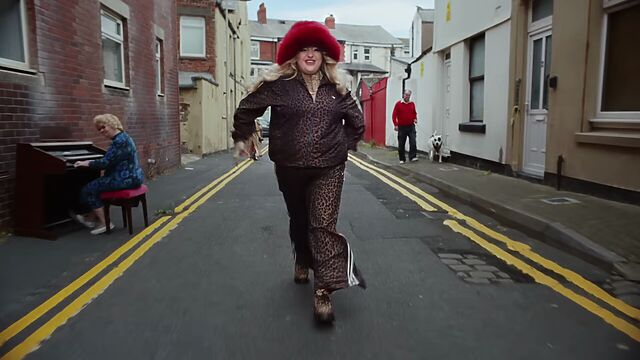 Now dressed in a leopardprint pantsuit and red furry hat, the singer walks down a narrow street, paved over cobblestones, while an organist plays on one side and a man walks his dog on the other.