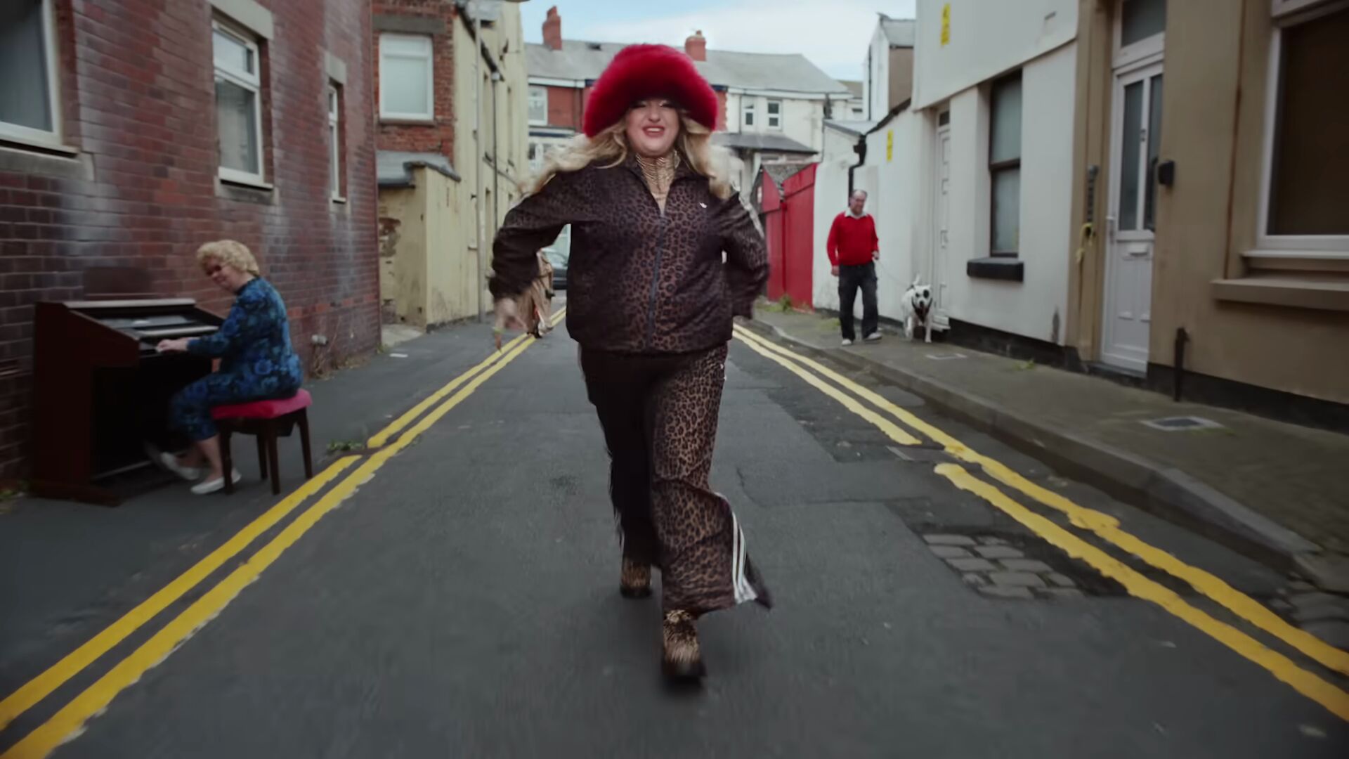 Full image of Now dressed in a leopardprint pantsuit and red furry hat, the singer walks down a narrow street, paved over cobblestones, while an organist plays on one side and a man walks his dog on the other.