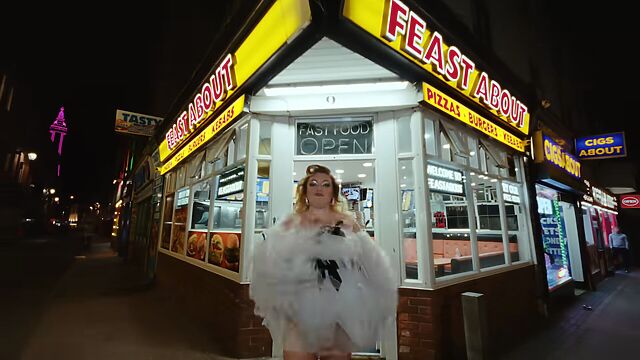 At nighttime, a woman in a wispy and feathery white dress stands outside Feast About Fast Food on a street corner, with illuminated Blackpool Tower just visible over the rooftops.