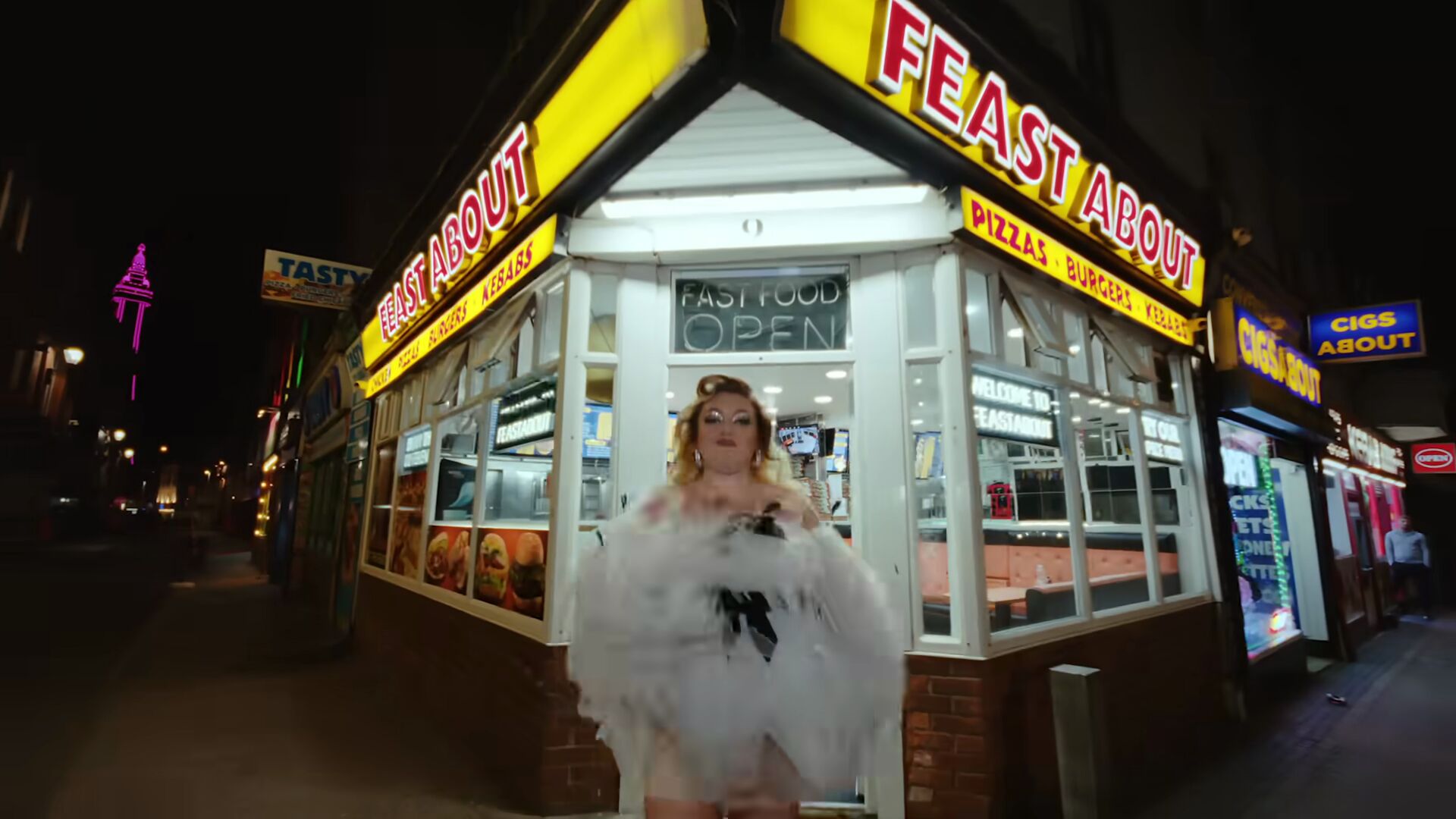 Full image of At nighttime, a woman in a wispy and feathery white dress stands outside Feast About Fast Food on a street corner, with illuminated Blackpool Tower just visible over the rooftops.
