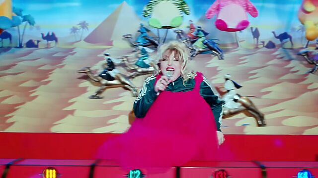 Model camels race across a desert at a sideshow stall, while a woman wearing a red strapped dress over a tracksuit sings energetically.
