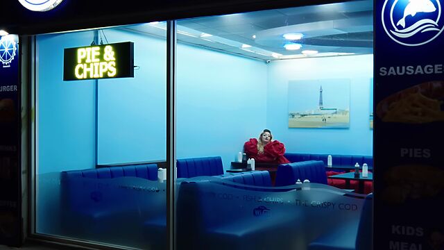 In a red poofy dress, a woman sits alone in a blue-lit fish and chip shop, as seen through the window, at night.