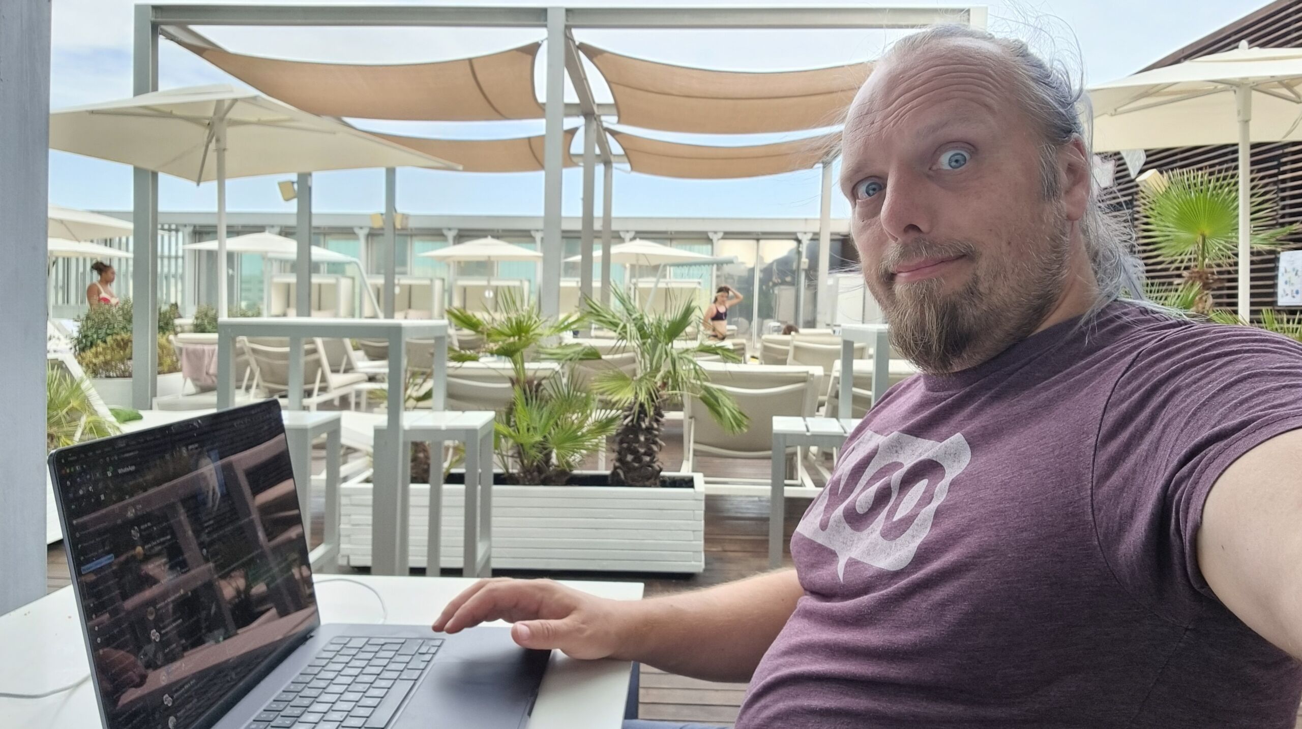 Full image of Dan, a white man with a goatee beard and a blue ponytail, sits at a poolside table in a hotel roof terrace, using his laptop alongside a pile of towels.
