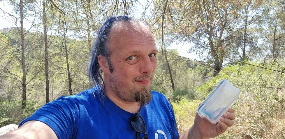 Dan, sweating, holds a medium-sized tupperware tub in a hilly forest.