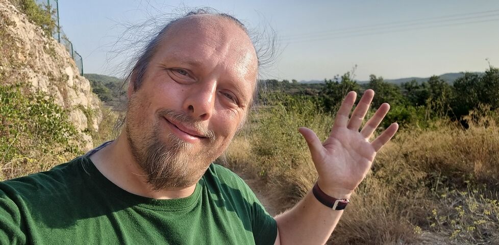 Dan, a white man in a green shirt, waves to the camera from a rural Spanish trail.