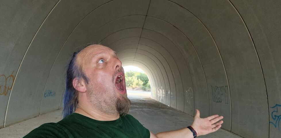 Dan, a white man with a blue ponytail, shouts upwards in a broad concrete tunnel.