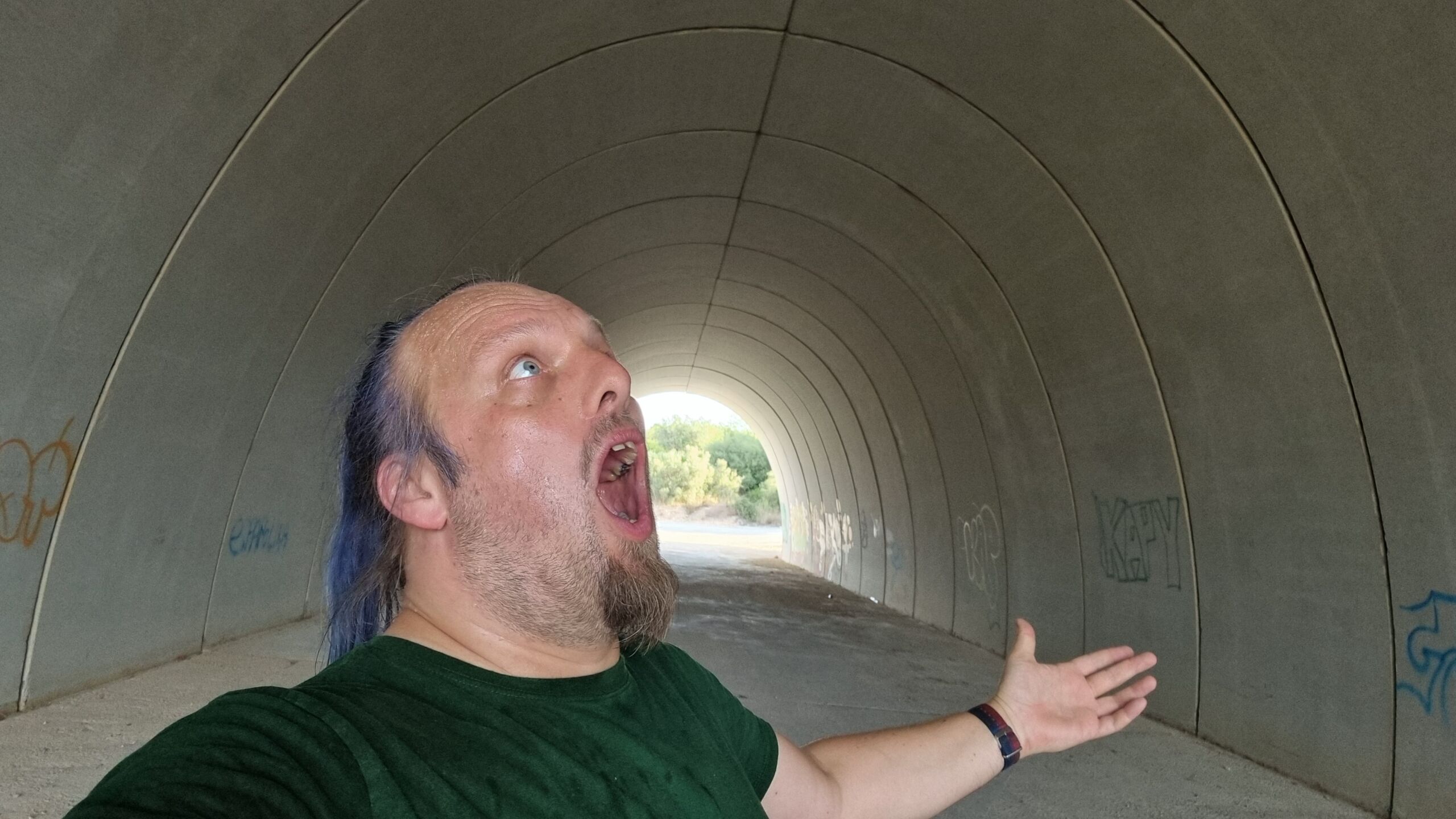 Full image of Dan, a white man with a blue ponytail, shouts upwards in a broad concrete tunnel.
