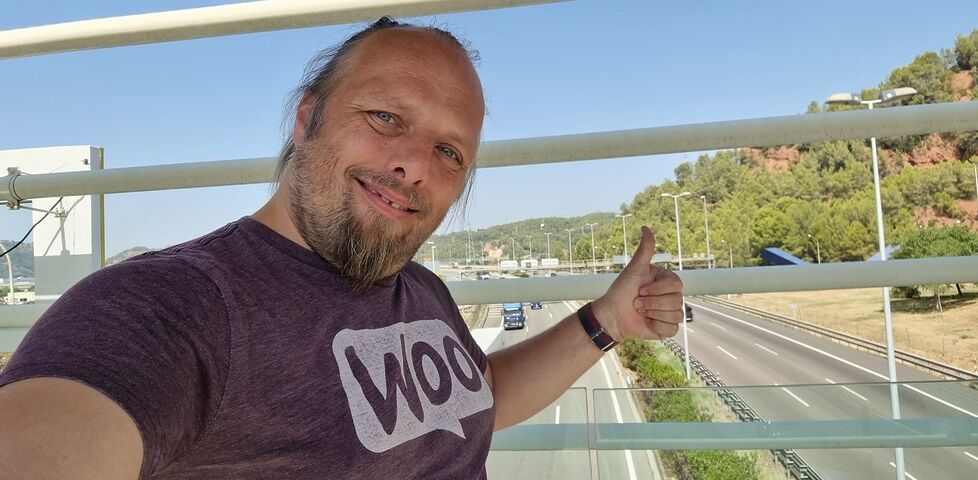On a semi-closed bridge over a six-lane highway, Dan, wearing a purple 'woo' t-shirt, throws a selfie camera a thumbs-up.