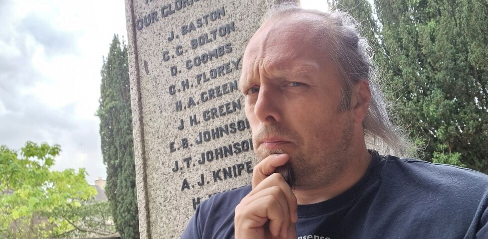 Dan, a white man with a ponytail and goatee beard, sits on a war memorial with a confused expression.