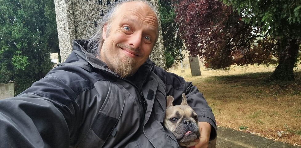 Dan, a white man with a goatee beard, sits on the steps of a rain-wettened village war memorial, hugging a fawn-coloured French Bulldog.
