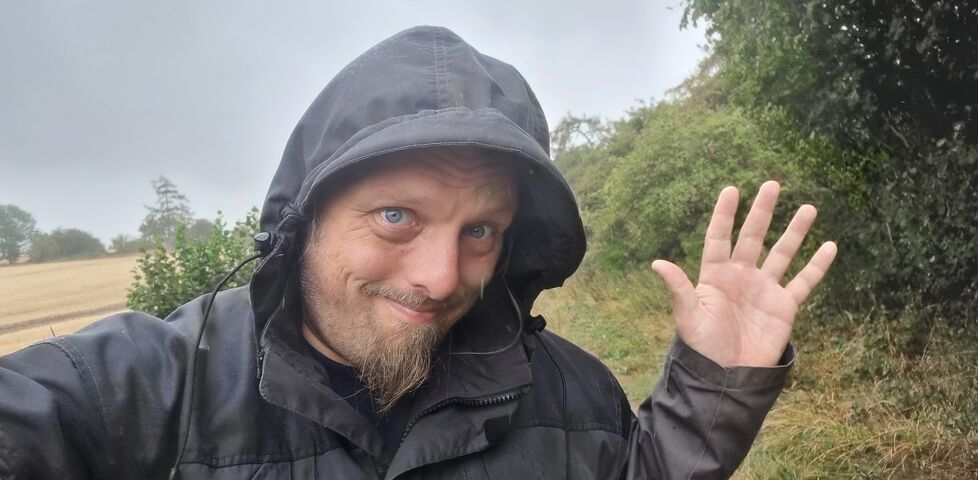 Dan, a white man with a goatee beard, wears a grey hooded jacket on a verdant rural footpath in moderate rain.
