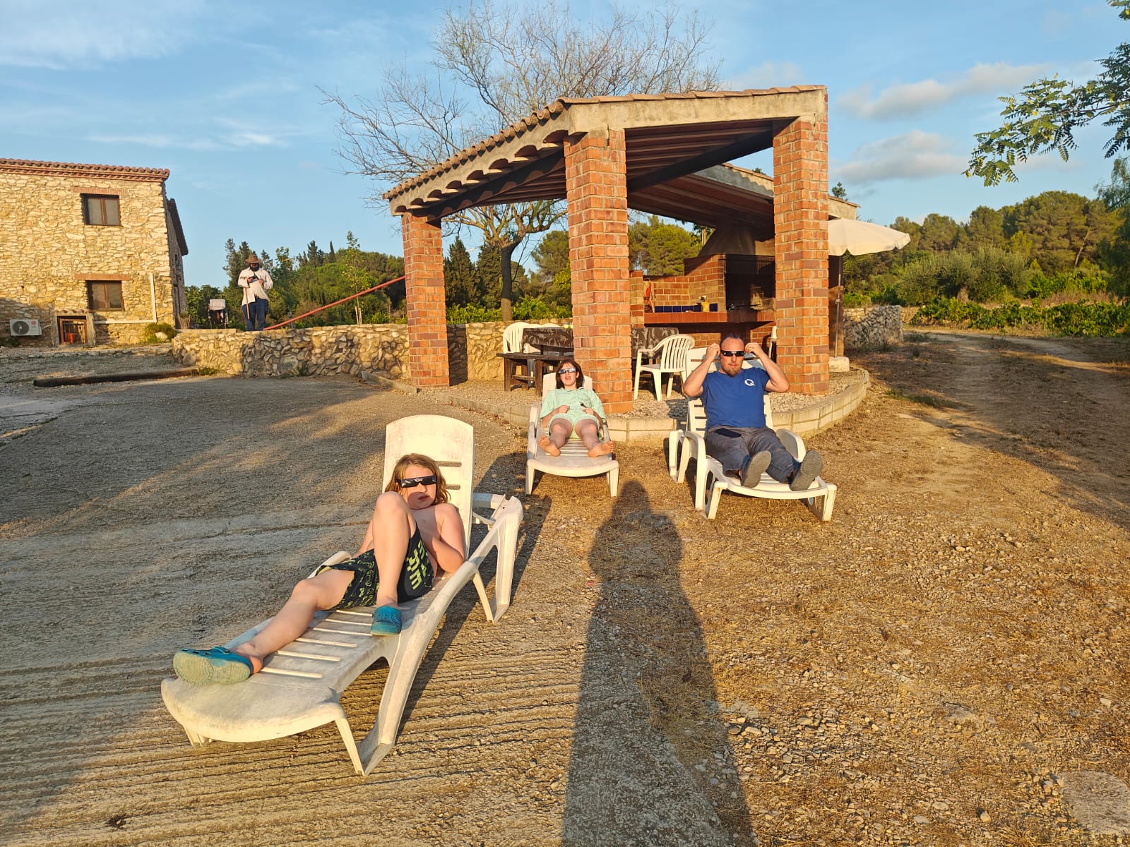 Full image of Dan sits on a sun lounger on a driveway, alongside a covered brick barbeque building. He's alongside two children doing the same, and all are wearing disposable eclipse glasses.