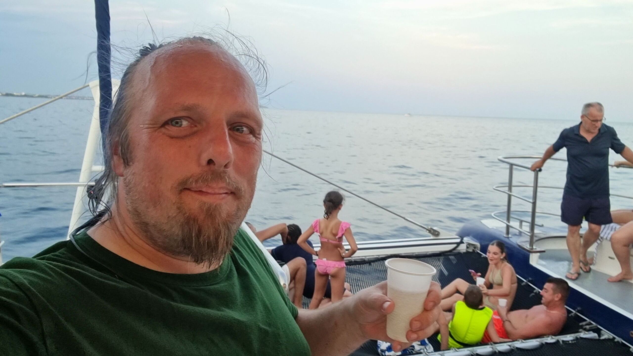 Full image of Dan, a white man, drips from a swim in the sea as he stands, holding a plastic cup of white wine, on the deck of a catamaran.