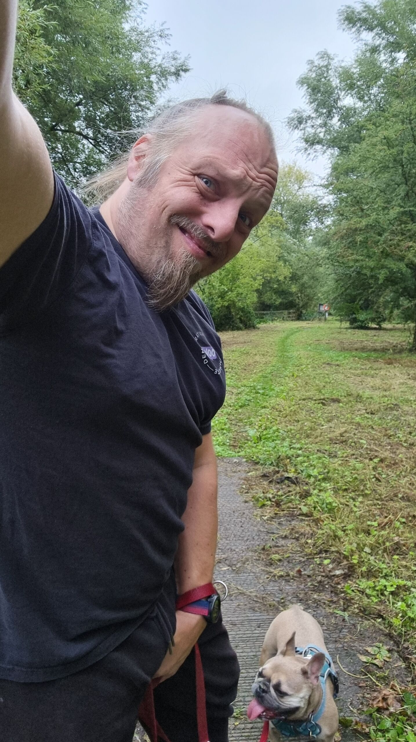 Full image of Dan, a white man with a goatee beard, grimaces as he holds his crotch, alongside a French Bulldog on a woodland footpath.