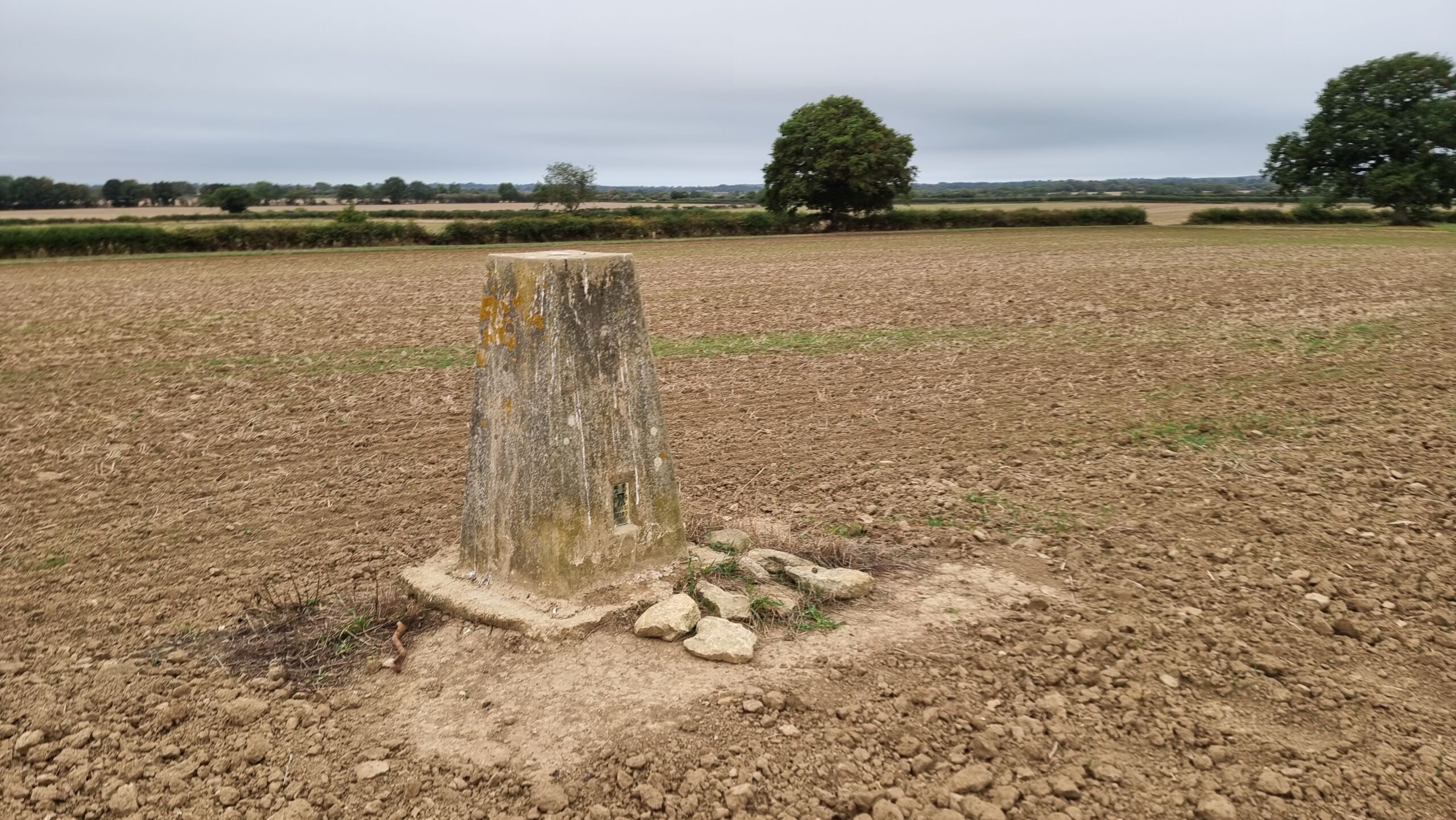 Full image of A concrete bollard style trigpoint stands in a bare field.