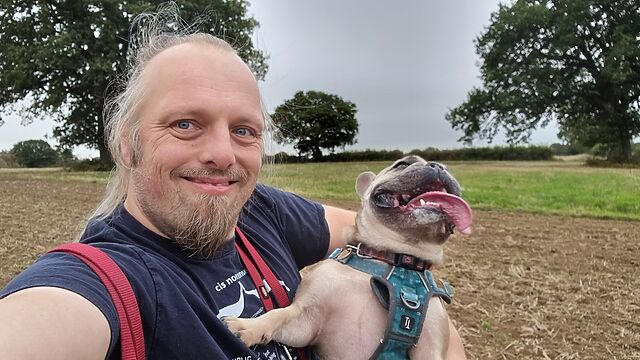 Dan, a white man with a goatee beard and ponytail, smiles for the camera while holding a French Bulldog whose tongue is lolling as she pants, in a field.