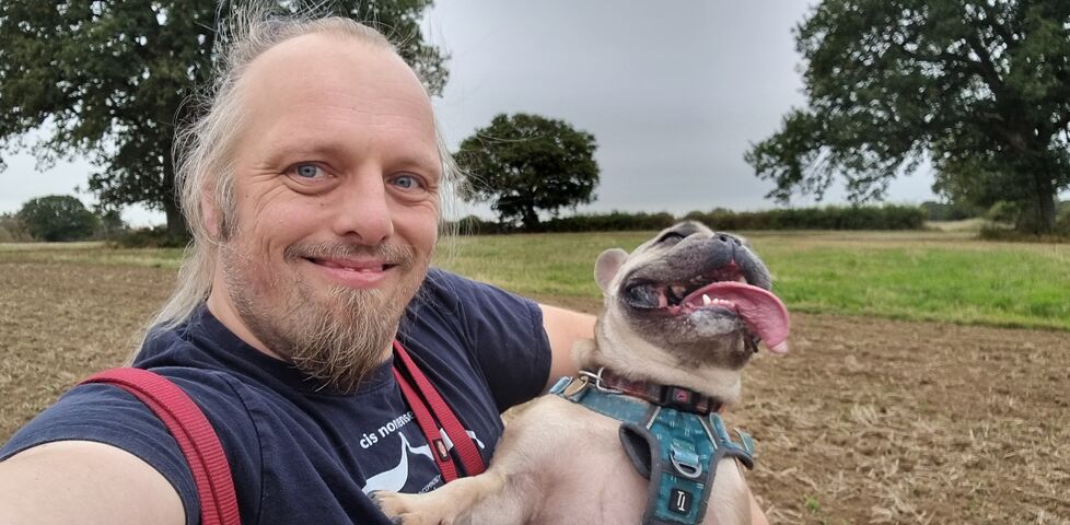 Dan, a white man with a goatee beard and ponytail, smiles for the camera while holding a French Bulldog whose tongue is lolling as she pants, in a field.