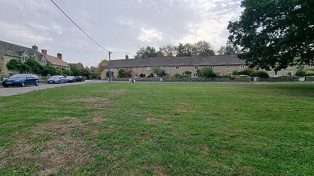 A mowed village green, surrounded by a road and a mixture of brick and stone houses.