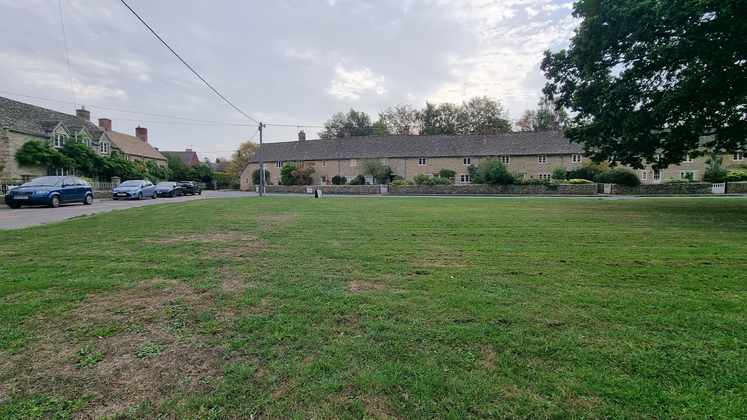 Full image of A mowed village green, surrounded by a road and a mixture of brick and stone houses.