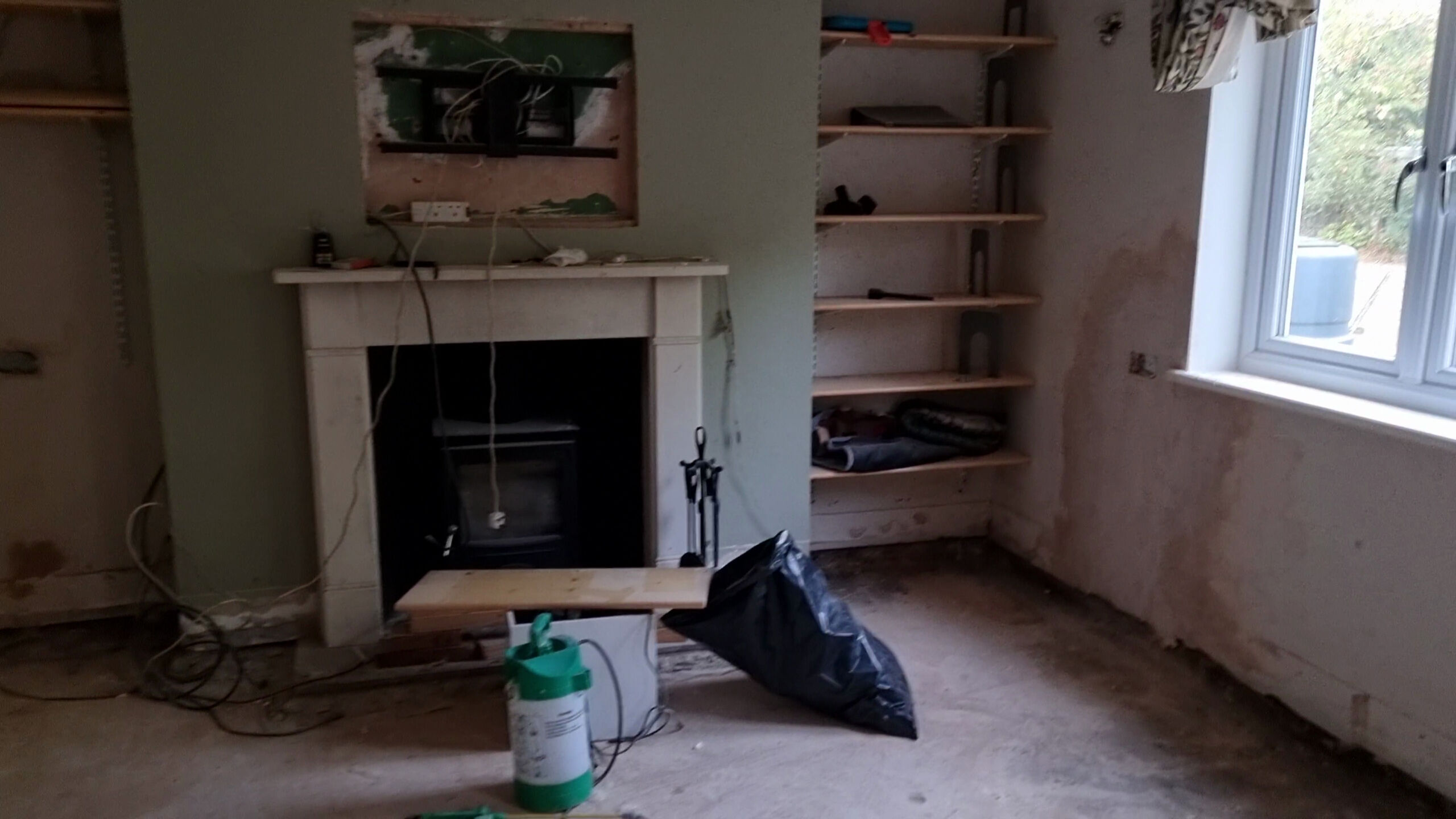 Full image of A dilapidated living room with damaged lower walls and missing flooring, filled with tools and bags of rubbish.