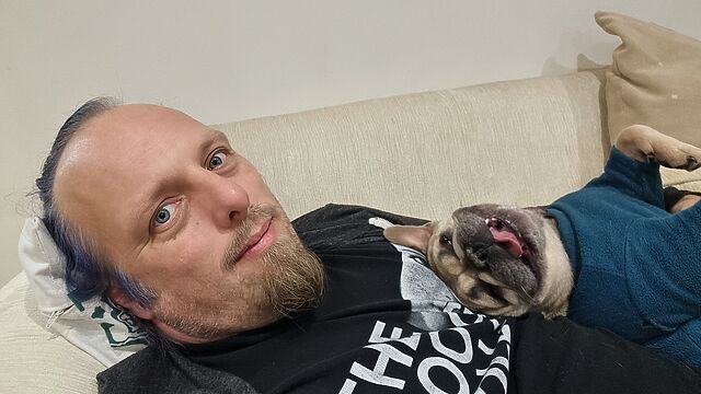 An inelegant looking French Bulldog in a teal jumper lies in her back, with her tongue out, on top of Dan.