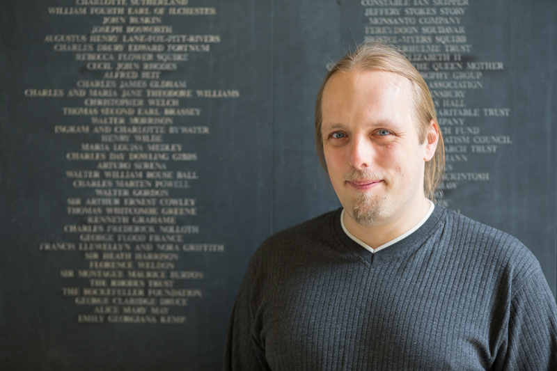 Full image of Dan, in a grey jumper, stands next to a stone plaque covered in names.