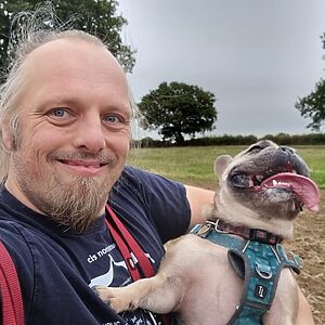 A French Bulldog with her tongue hanging out is lifted by Dan.