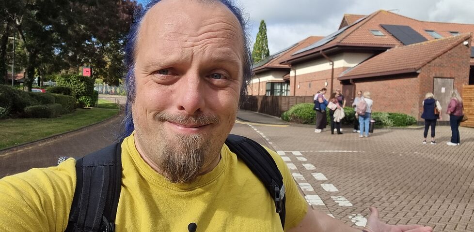 Dan, a white man with a goatee beard and a blue ponytail, walks away from a crowd gathered on the road outside a hotel building.