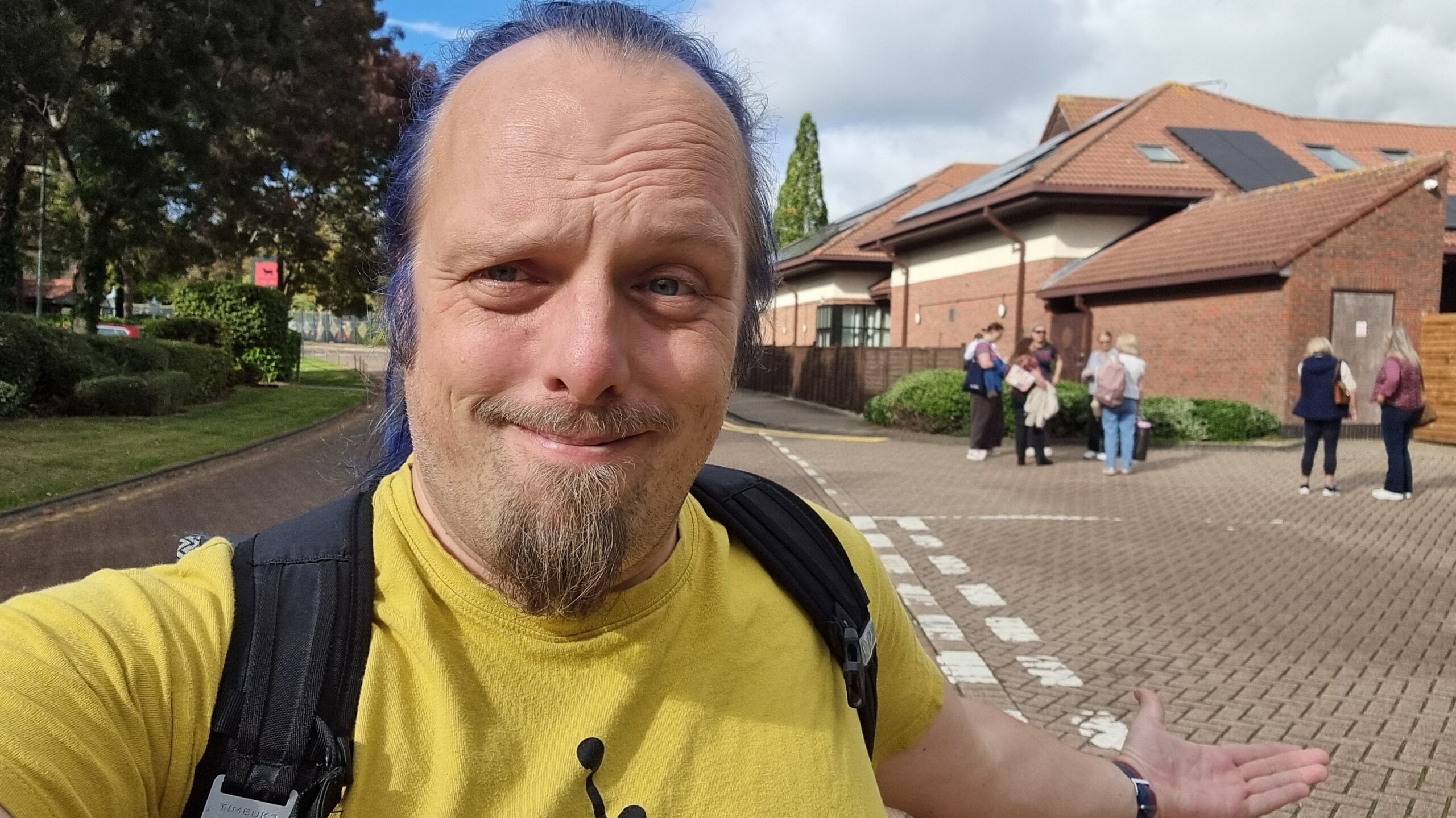 Full image of Dan, a white man with a goatee beard and a blue ponytail, walks away from a crowd gathered on the road outside a hotel building.
