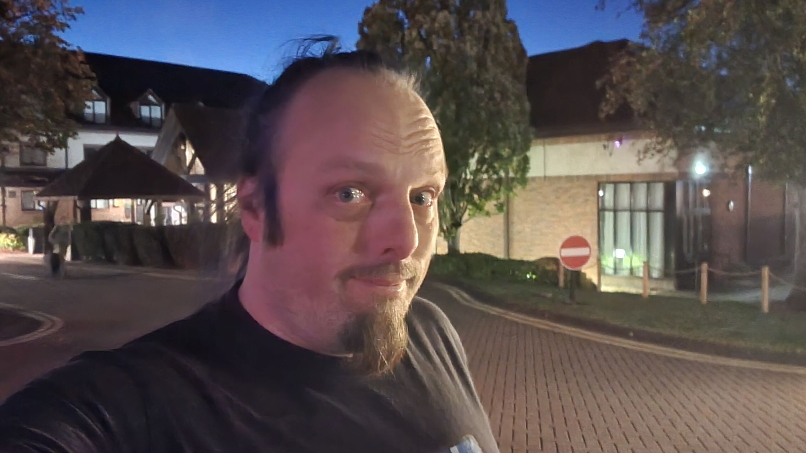 Full image of Dan, a white man with a blue ponytail and a goatee beard, stands in the dark in the car park of a hotel building.