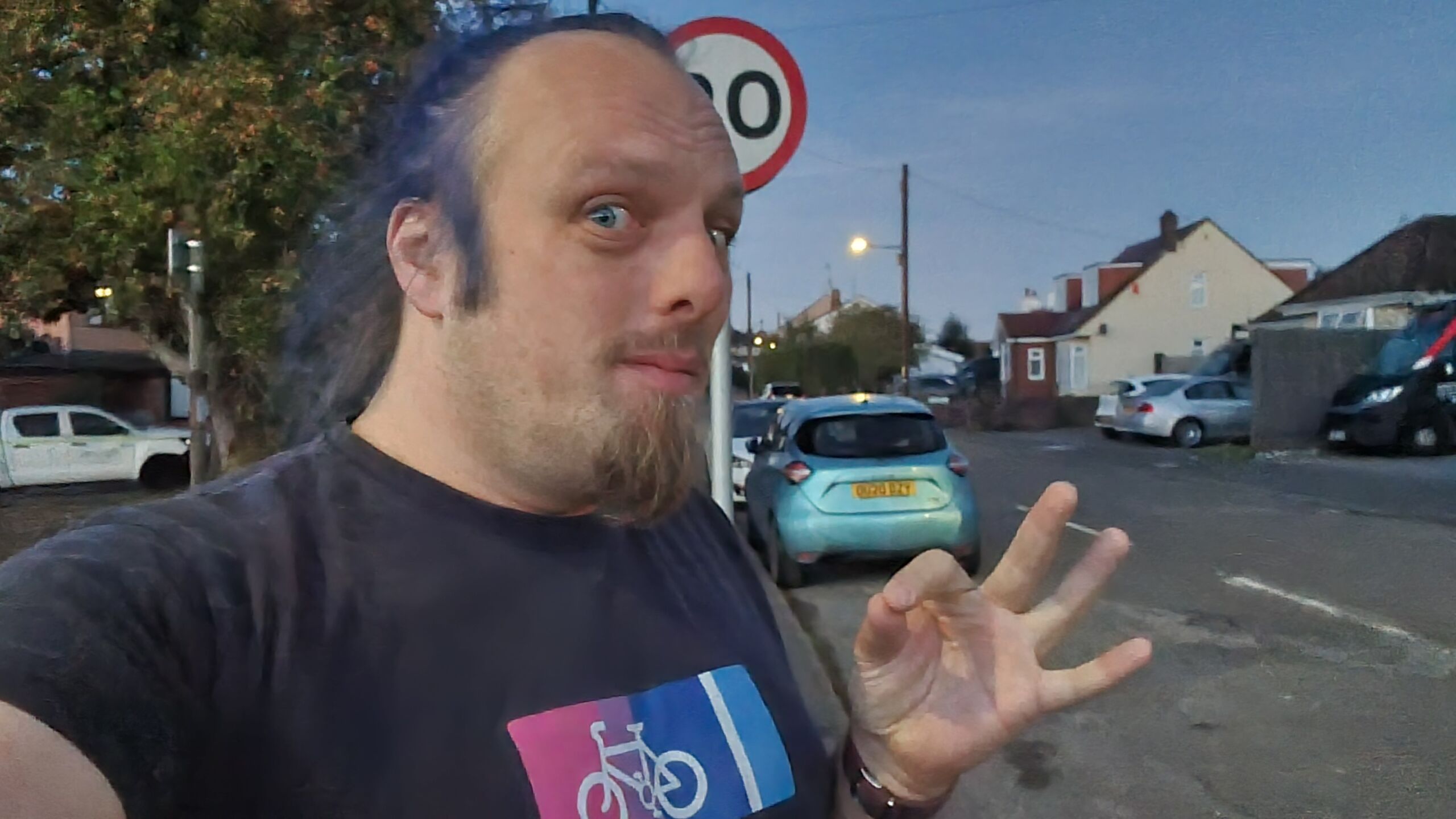 Full image of Dan, now seen to be wearing a "bi cycle lane" t-shirt in bi pride colours, throws an "okay" sign in front of a parked car.