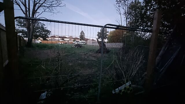 Through a tall wire fence a grassy area with rubble and a prominent tree stump.