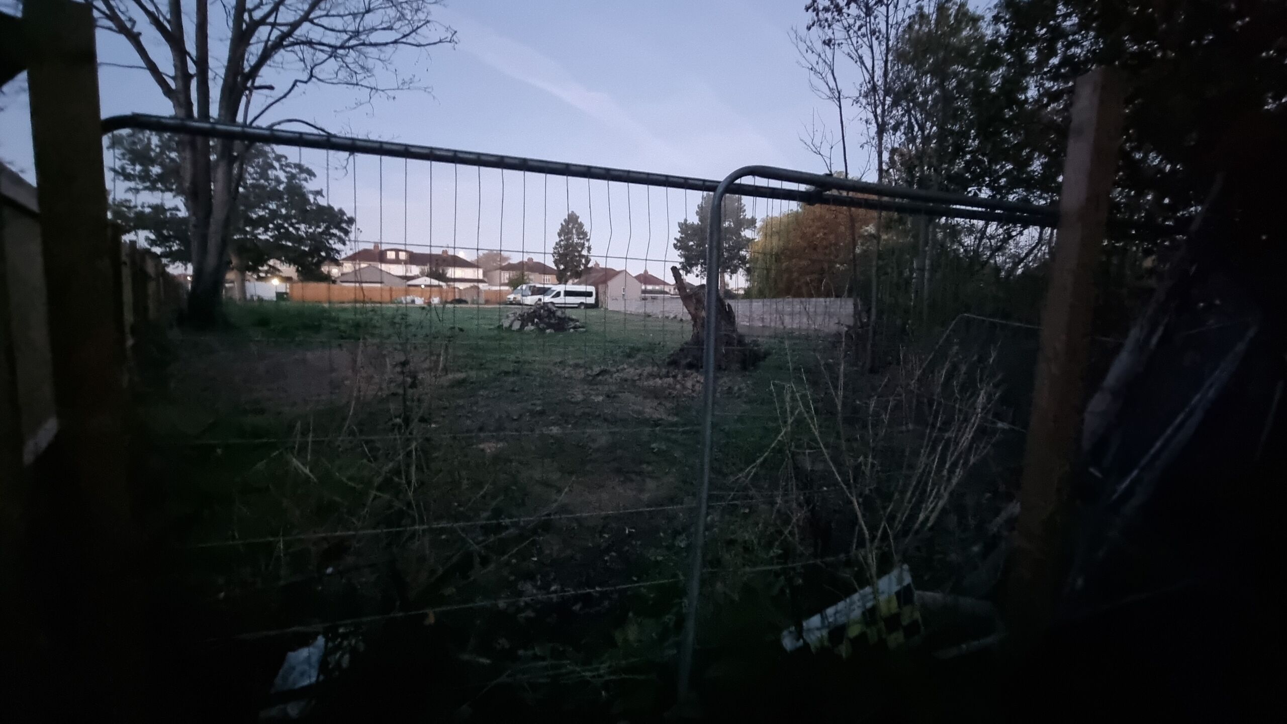 Full image of Through a tall wire fence a grassy area with rubble and a prominent tree stump.