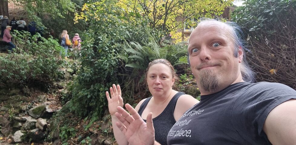 Dan and Ruth sit on a bench alongside a verdant spring pool, as spectators cheer on runners in the background.