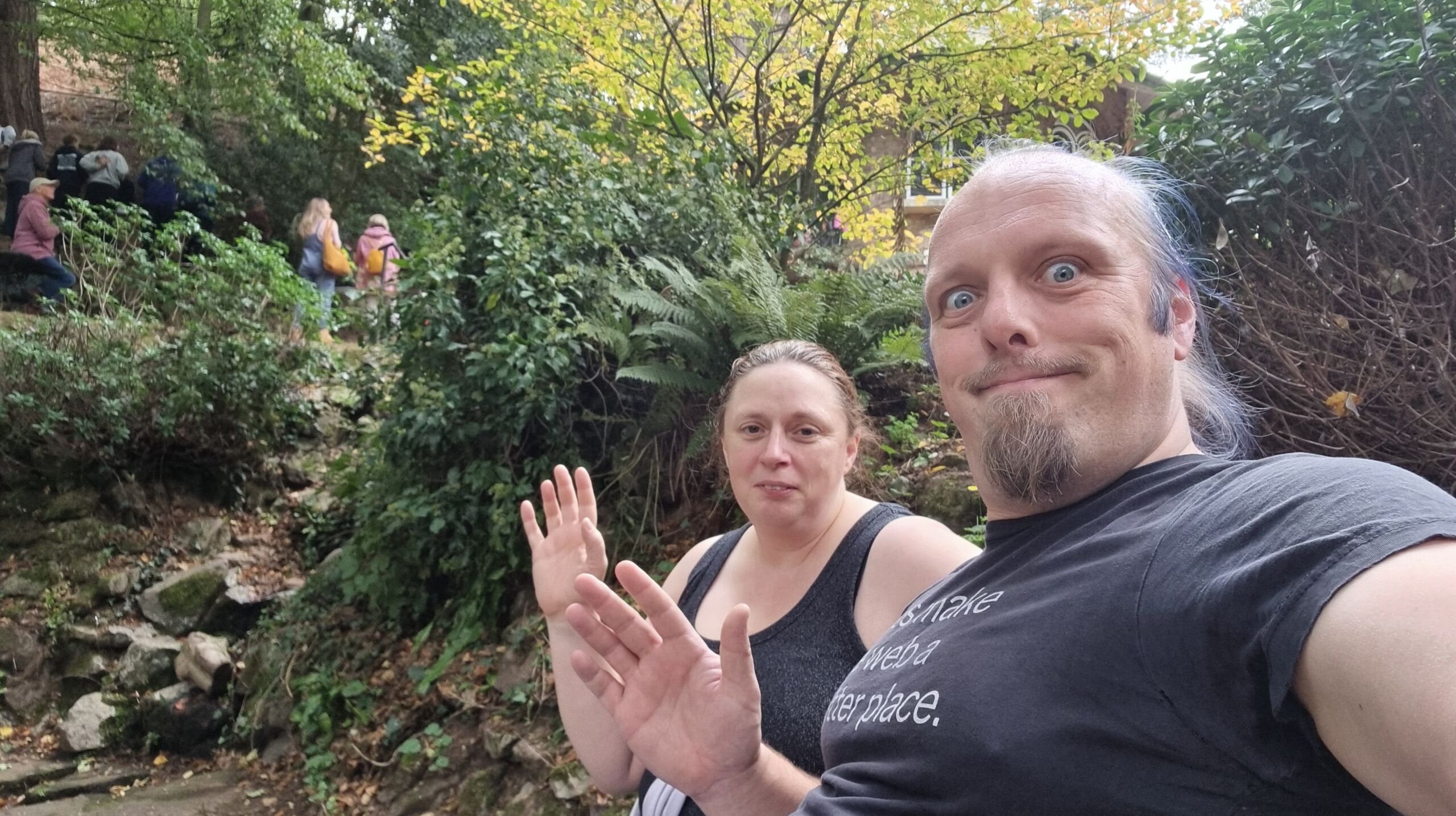 Full image of Dan and Ruth sit on a bench alongside a verdant spring pool, as spectators cheer on runners in the background.