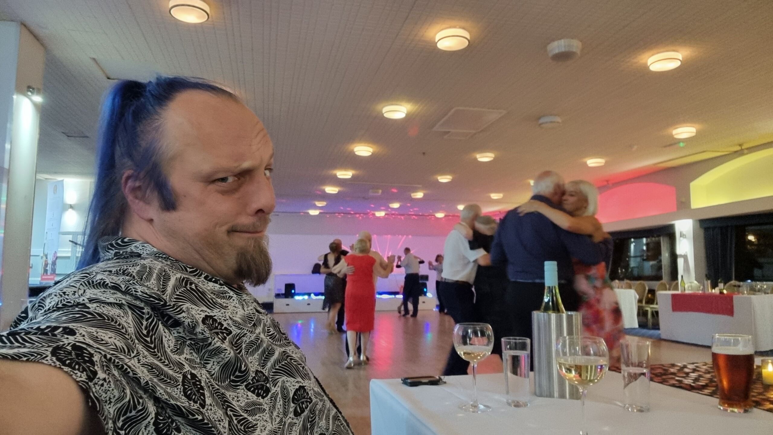 Full image of Dan, a white man with a goatee beard and blue ponytail, wearing a monochrome leaf-pattered shirt, sits at a table full of beer and wine, squinting quizzically at the camera while dancers shuffle around a dance floor behind him.