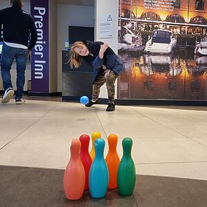 In the lobby of a Premier Inn, a child bowls with a plastic ball and pins.