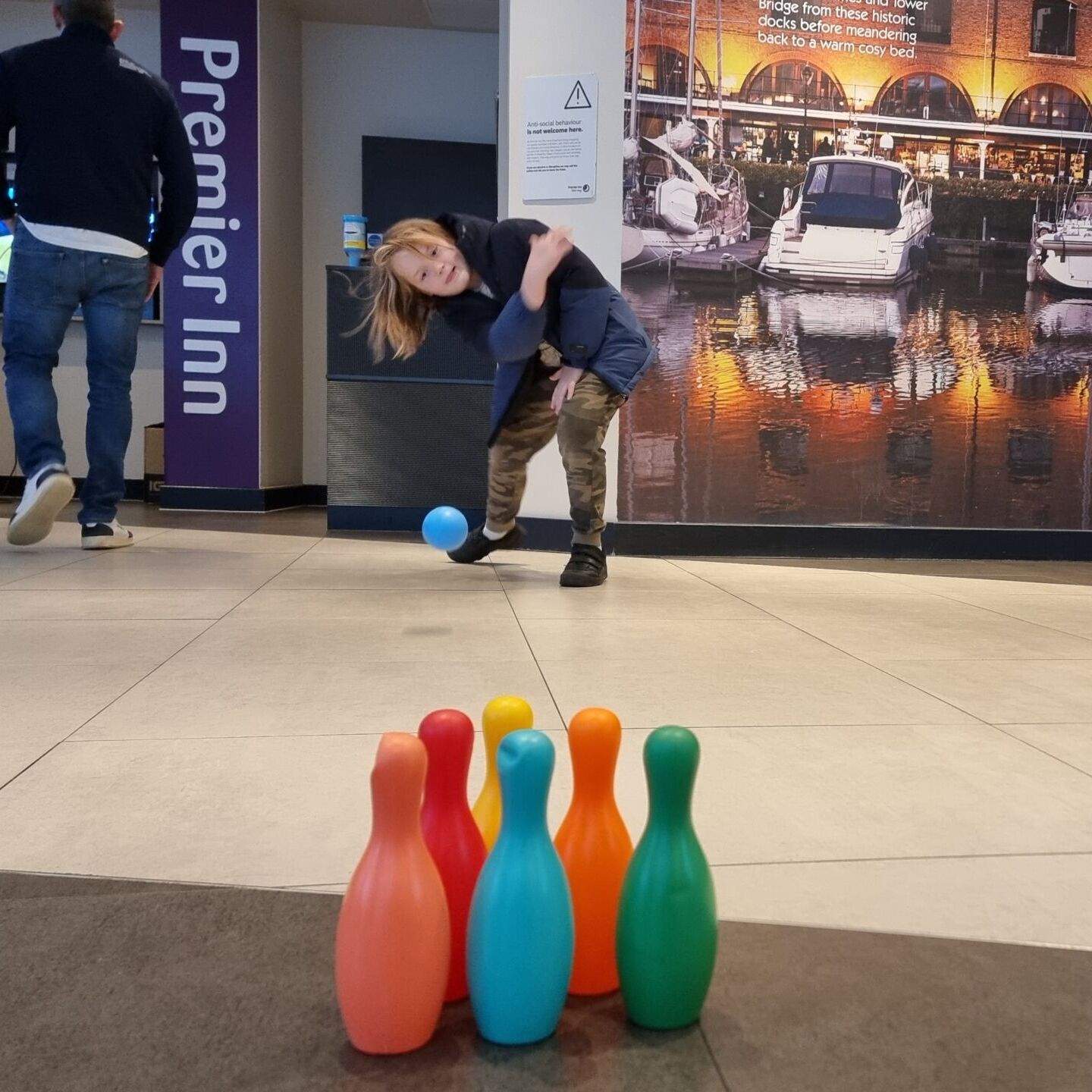 Full image of In the lobby of a Premier Inn, a child bowls with a plastic ball and pins.