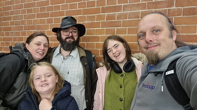 A smiling family including Dan pose for a group selfie in front of a red brick wall.
