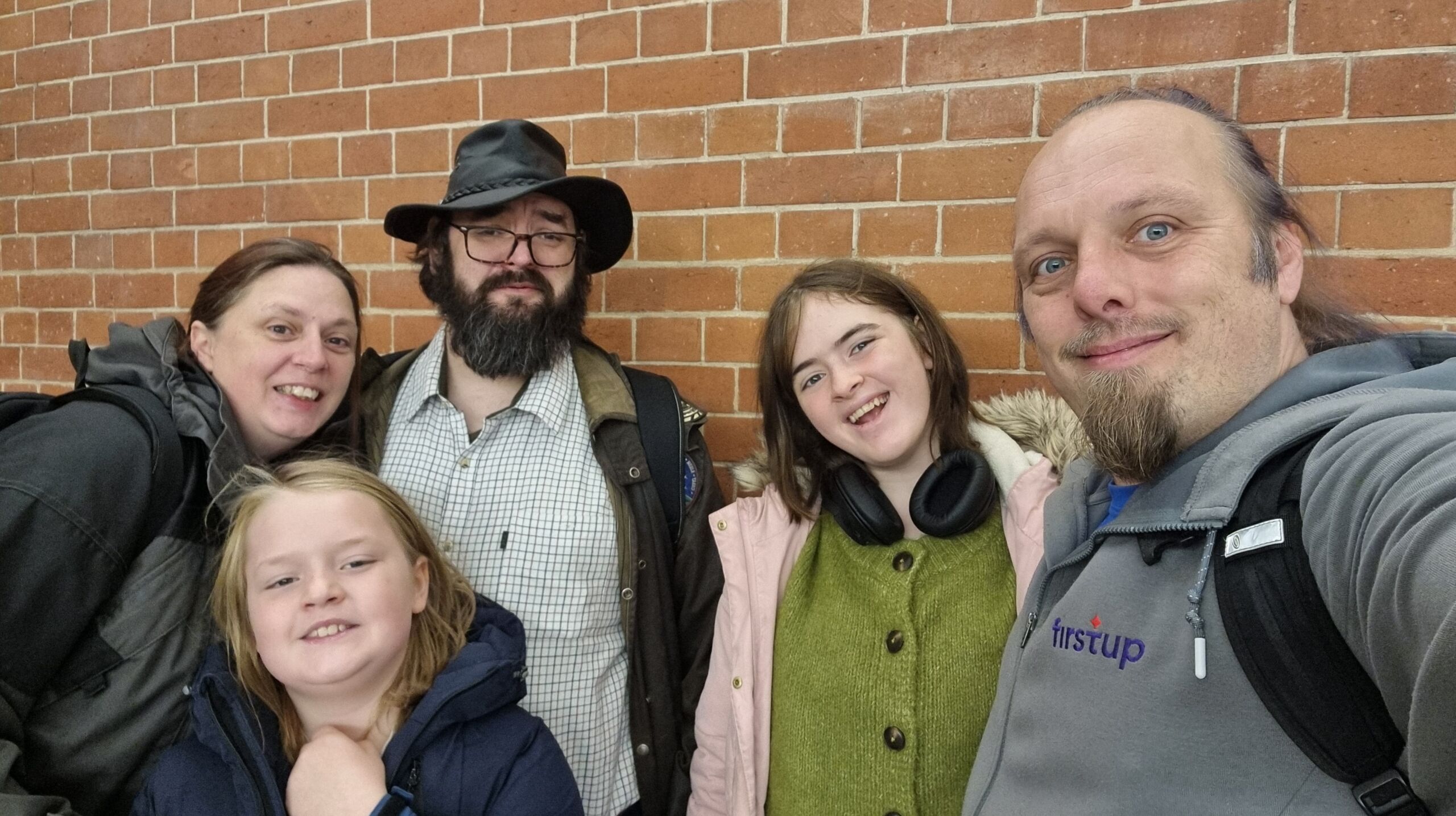Full image of A smiling family including Dan pose for a group selfie in front of a red brick wall.