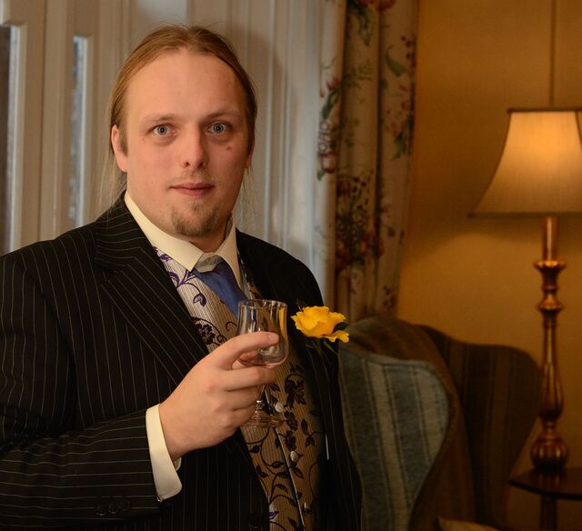 Dan, aged around 30, wearing a suit, waistcoat, buttonhole and cravat, holds a small glass in a traditionally but attractively-decorated hotel.