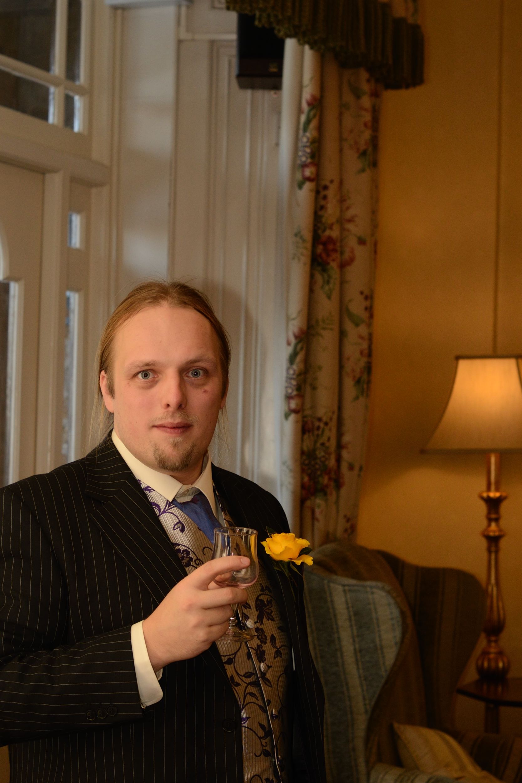 Full image of Dan, aged around 30, wearing a suit, waistcoat, buttonhole and cravat, holds a small glass in a traditionally but attractively-decorated hotel.